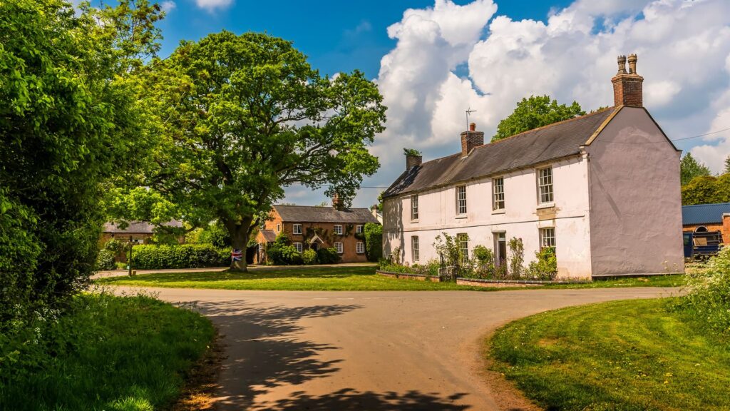 Traditional white cottages beside a village green, framed by mature trees under a bright blue sky